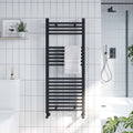 Black towel radiator mounted on a tiled bathroom wall with a white bathtub and shower in the background.